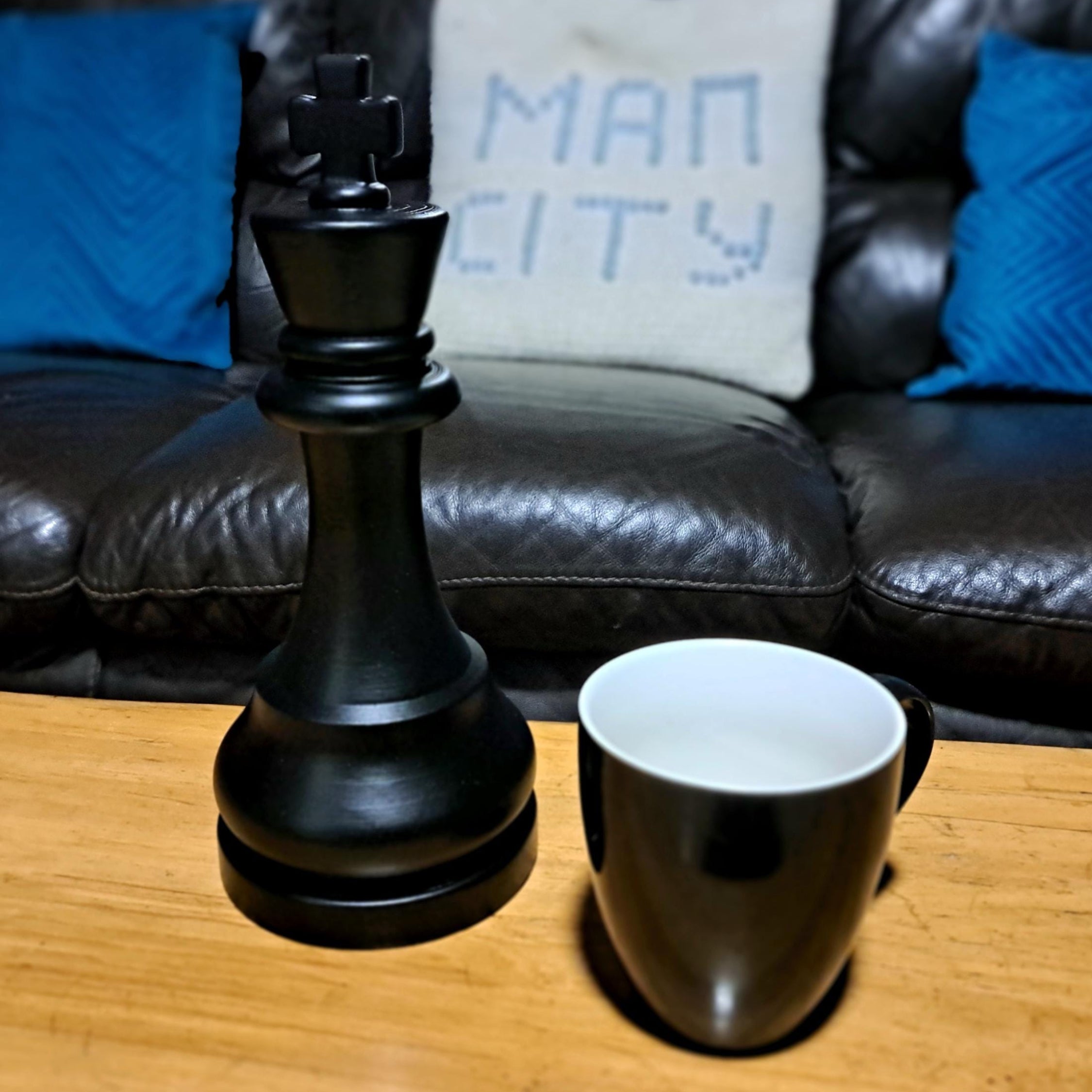 Large black king chess piece next to a black mug on a wooden table with a sofa in the background.