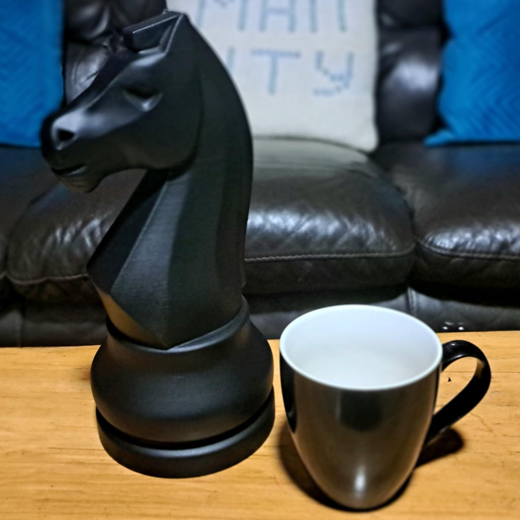Large black knight chess piece resembling a horse next to a white mug on a wooden surface with a couch in the background.