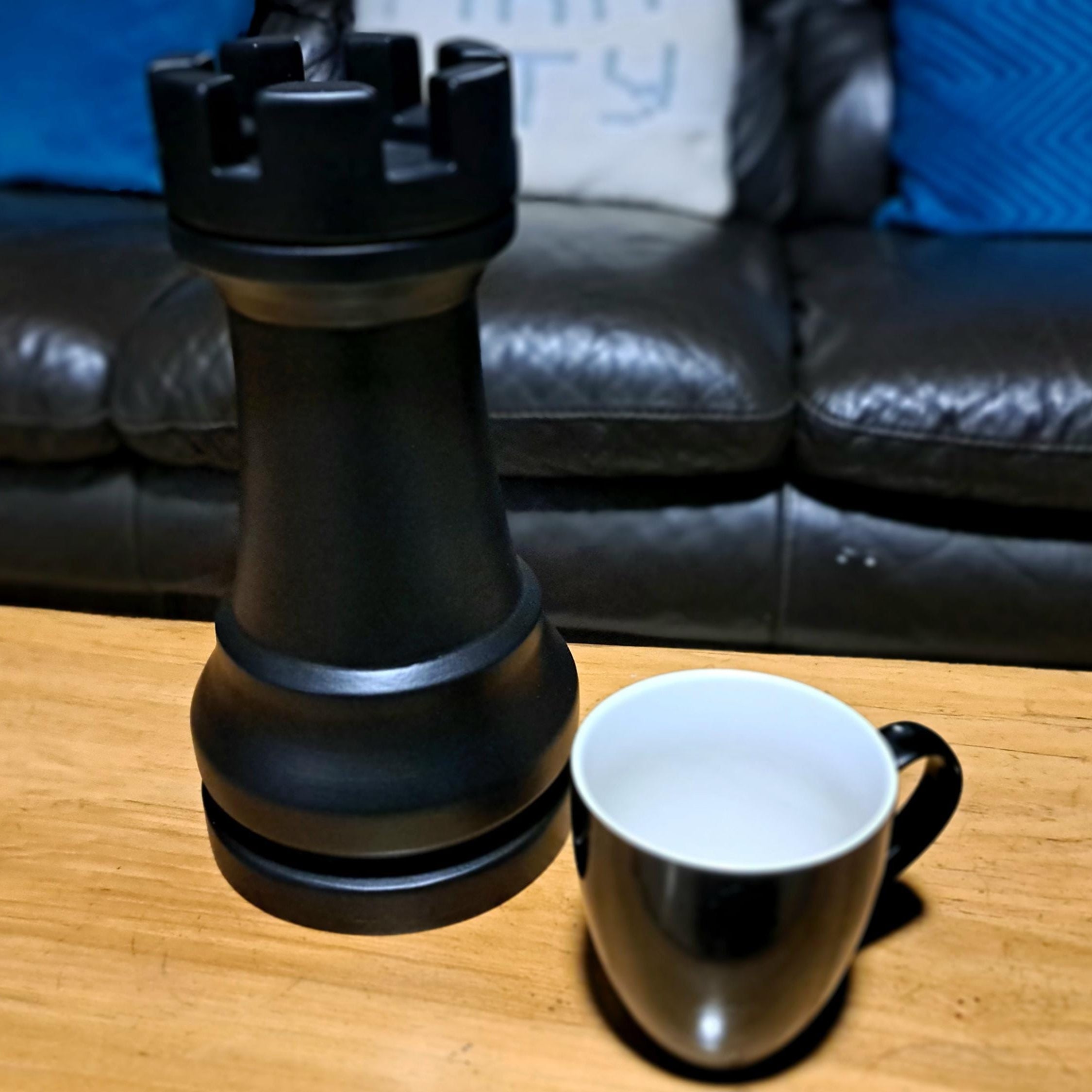 Large black rook chess piece next to a white mug on a wooden table with a couch in the background.