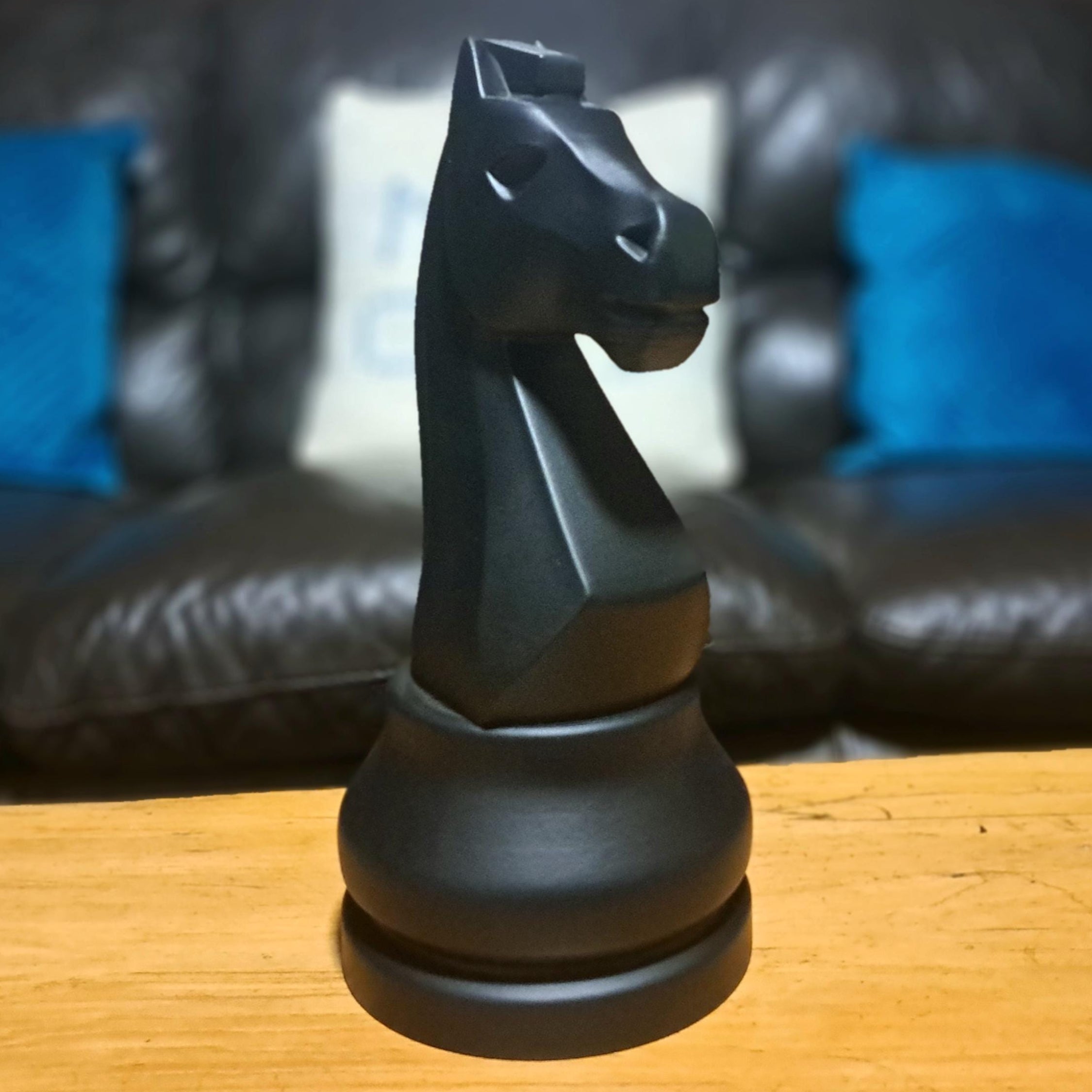 Large black knight chess piece on a wooden surface with a blurred background of a couch and pillows.