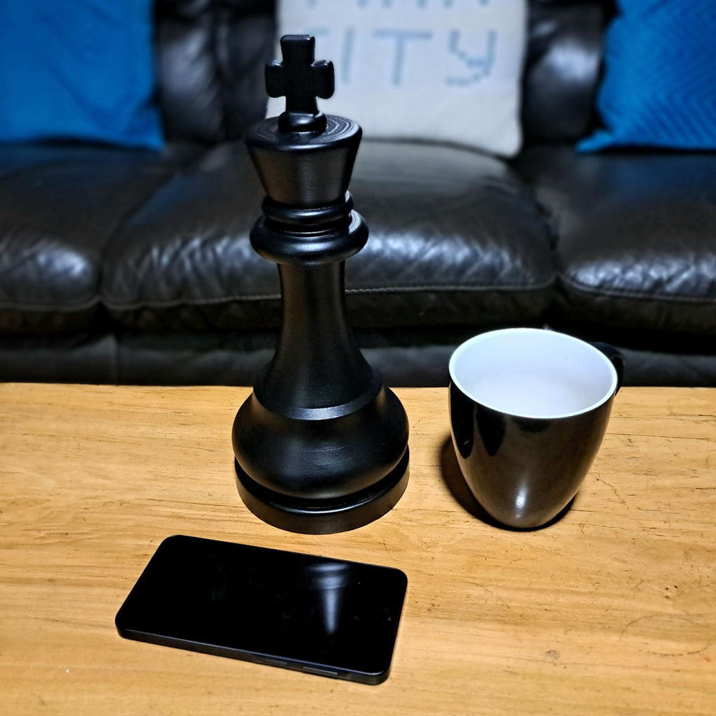 Large black king chess piece next to a black mug on a wooden table with a couch in the background.