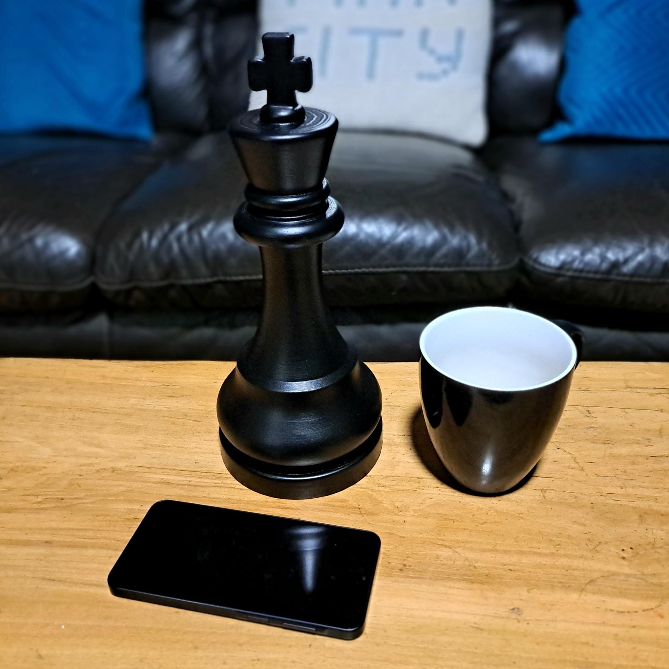 Large black king chess piece next to a black mug on a wooden table with a couch in the background.