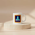 A white ceramic mug with a custom graphic print featuring a blue background, a teal silhouette, and the word 'BALDI' in white, set on a round beige stone pedestal against a warm beige background.