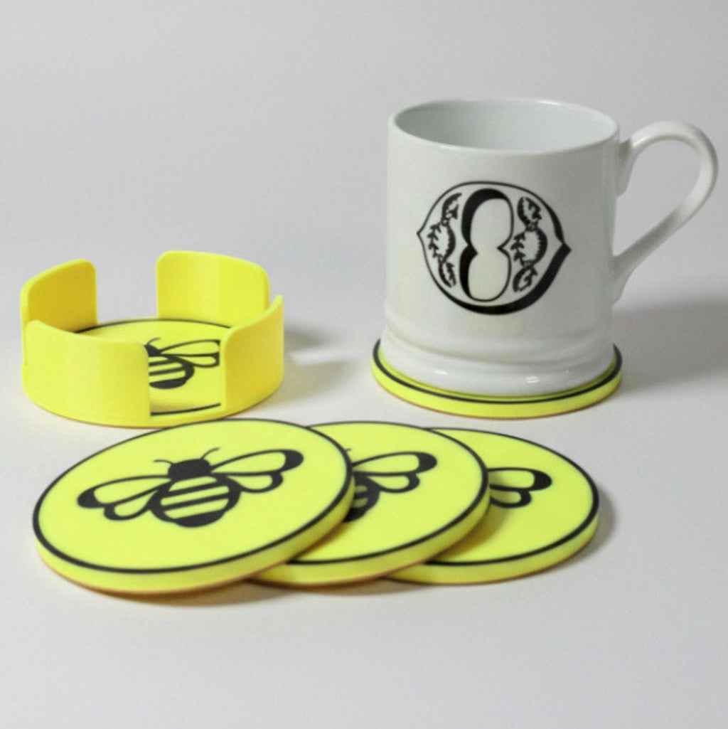 A set of bright yellow coasters and a matching holder with black bee graphics, shown with a white ceramic mug on a light gray background.