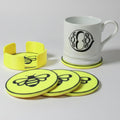 A set of bright yellow coasters and a matching holder with black bee graphics, shown with a white ceramic mug on a light gray background.