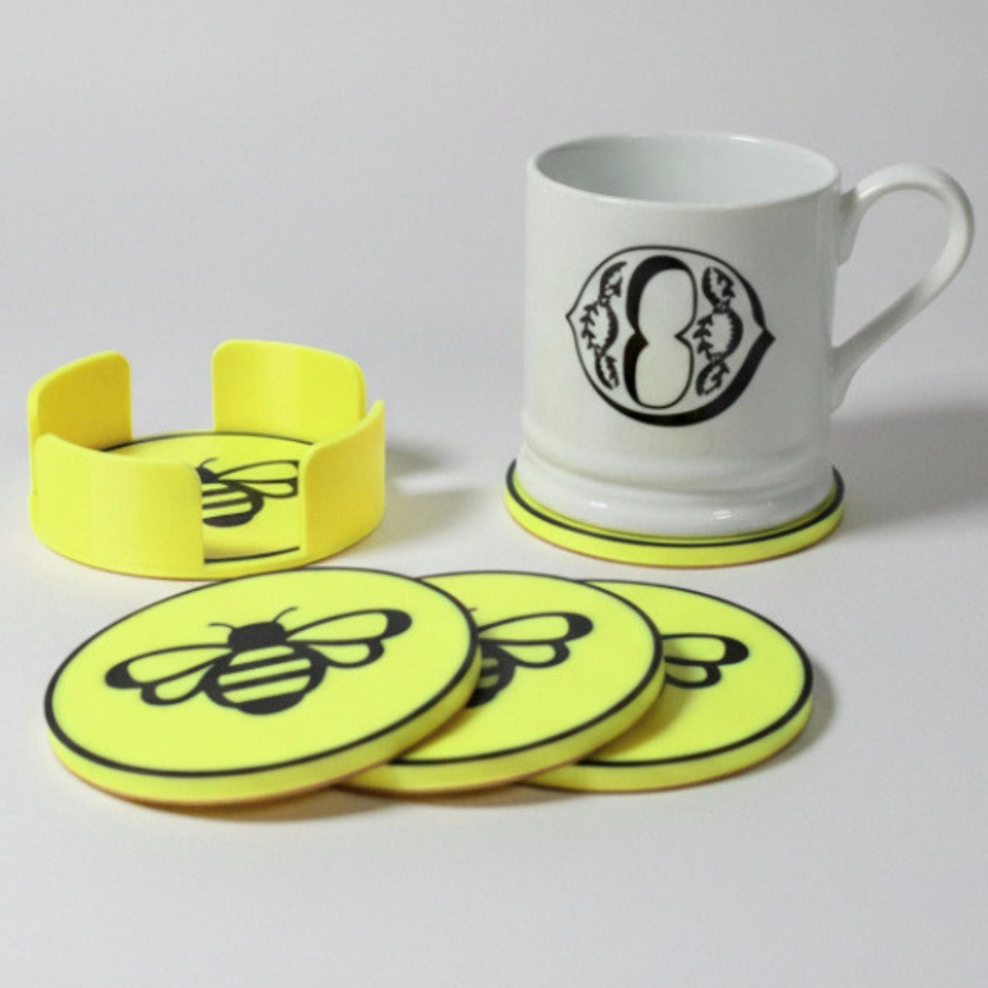 A set of bright yellow coasters and a matching holder with black bee graphics, shown with a white ceramic mug on a light gray background.
