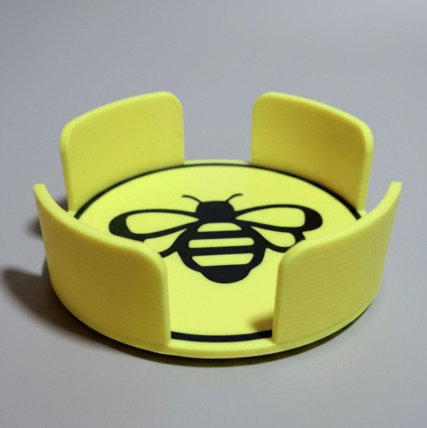 A bright yellow circular coaster holder with a black printed bee design in the center and four raised side grips, shown on a neutral gray background.