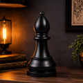 A matte black chess-piece sits on a wooden table in a warmly lit room setting with a lit lamp, books, and a plant in the background.