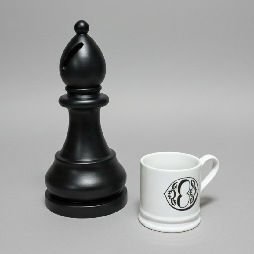 A black bishop chess-piece-shaped object standing next to a white ceramic mug with a black monogram logo, photographed on a neutral gray studio background.