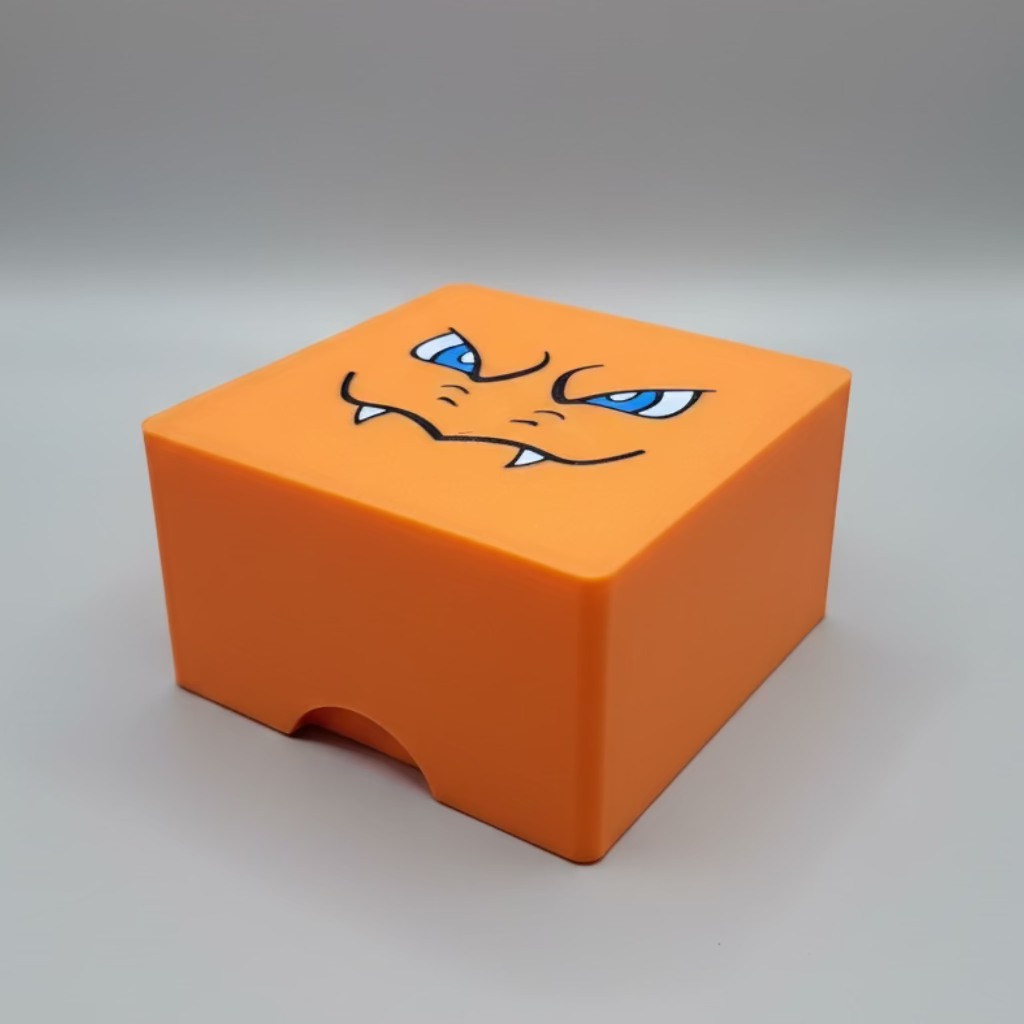 An orange cube-shaped deck box, featuring a printed charazard face on the top lid with blue eyes and small white fangs, set against a neutral gray background.