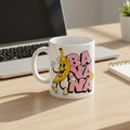 A white ceramic coffee mug with a cartoon banana character and the word 'BANANA' in pink bubble letters, sitting on a light wood desk next to a laptop.