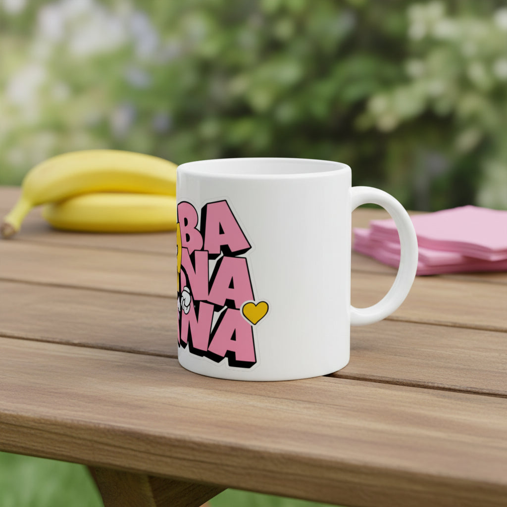 A white ceramic mug featuring a graphic print of the word 'BANANA' in bold pink letters with black outlines and a small yellow heart accent, shown on a clean white background.