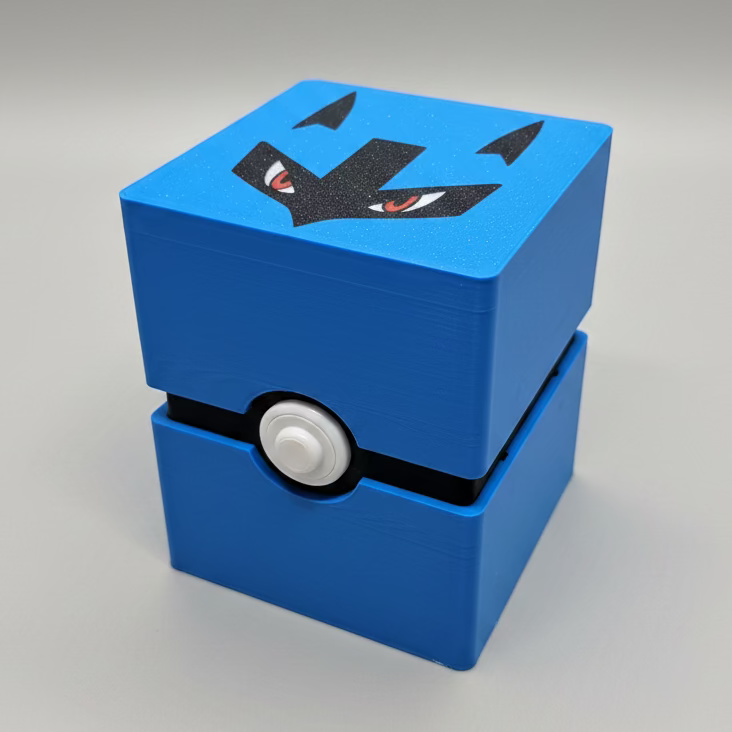 An angled, close-up view of a closed 3D-printed Pokemon TCG deck box. The box is vibrant blue with a black horizontal band around the middle, featuring a white Pokéball-style button in the center. The top lid displays Lucario's signature black mask and red eyes.