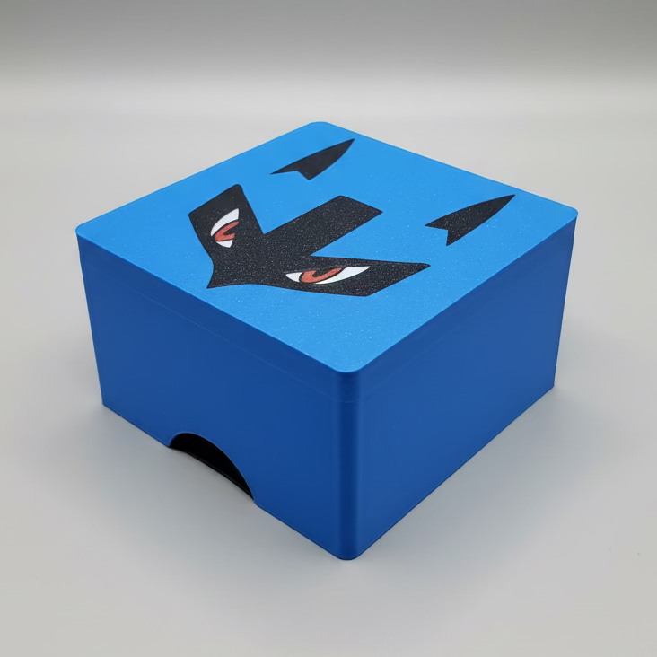 A square, bright blue 3D-printed lid for a Pokemon TCG deck box. The smooth top surface features a bold, black graphic of Lucario’s face details, including its sharp black mask and narrowing red eyes. The bottom edge of the blue lid has a semi-circular cutout for easy opening.