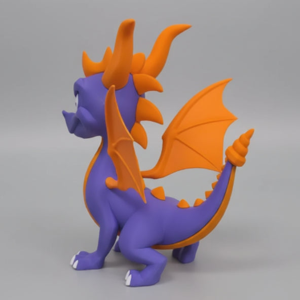 A purple and orange spyro the dragon figurine standing against a light gray wall. The dragon has orange horns, wings, and spikes, with white claws on its feet.