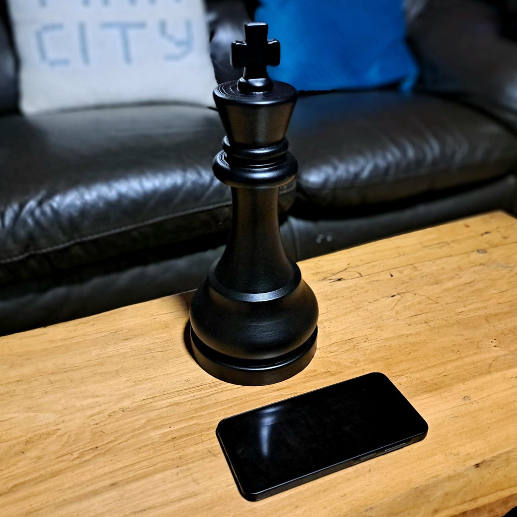 Large black chess piece on a wooden table with a phone below it, against a blurred background of a couch and pillow.