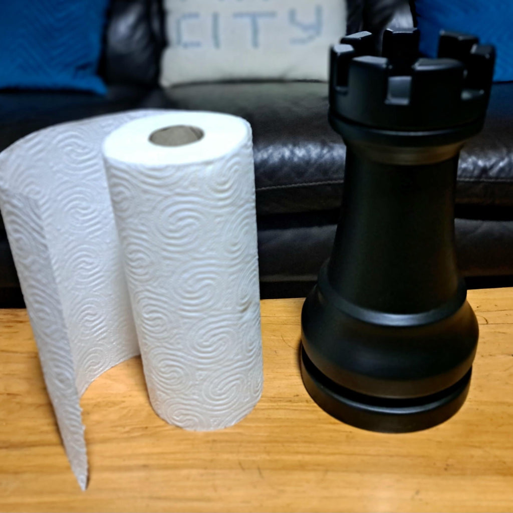 Black chess piece next to a roll of paper towels on a wooden table with a sofa in the background.