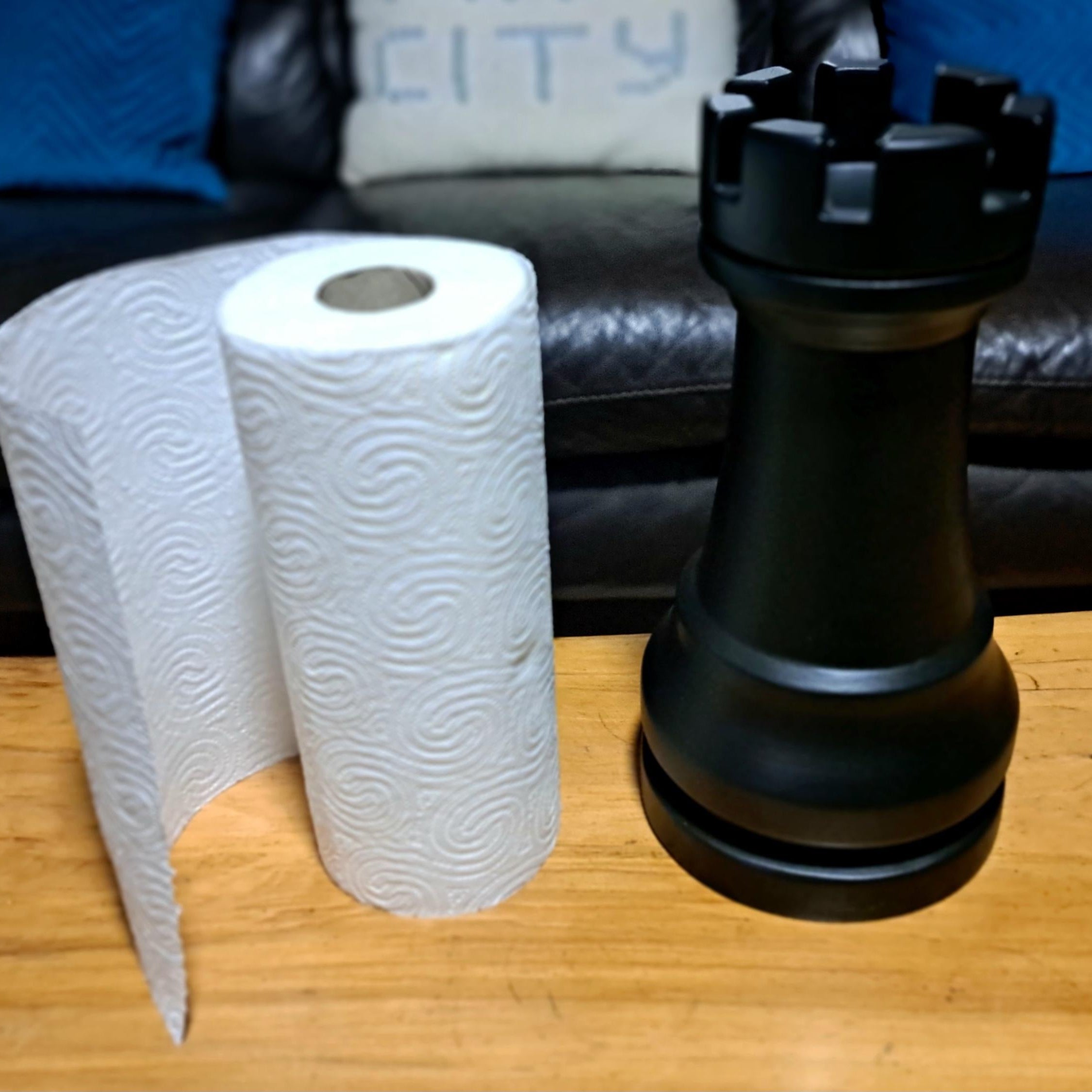 Black chess piece next to a roll of paper towels on a wooden table with a sofa in the background.