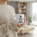Woman in a cream knit jumper holding a steaming Keep Calm and Drink Coffee white ceramic mug in a bright living roomsetting.